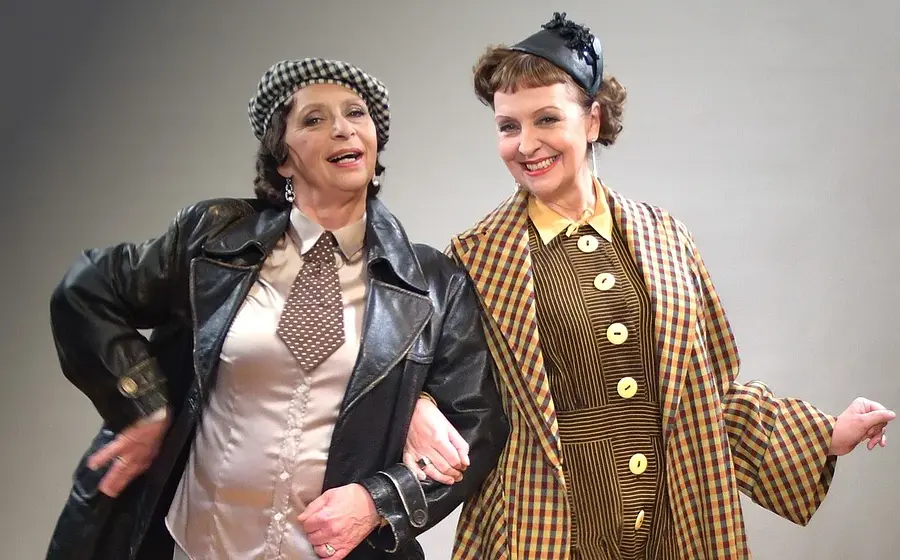 Susanne Brantl und Conny Glogger in historisch inspirierter Kleidung auf einer Bühne. Eine Frau trägt Lederjacke, Krawatte und karierte Mütze, die andere einen karierten Mantel mit schwarzem Hut. Beide haken sich unter und lächeln.