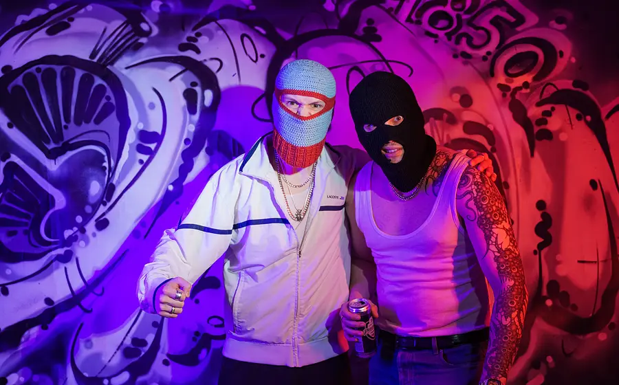 Zwei maskierte Personen vor Graffiti‑Wand, Neonlicht in Blau und Pink, halten Sprühdose.