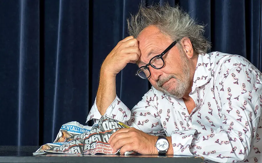 Urban Priol dien Kopf auf die Hand gestützt in gemustertem Hemd vor dunkelblauem Vorhang
