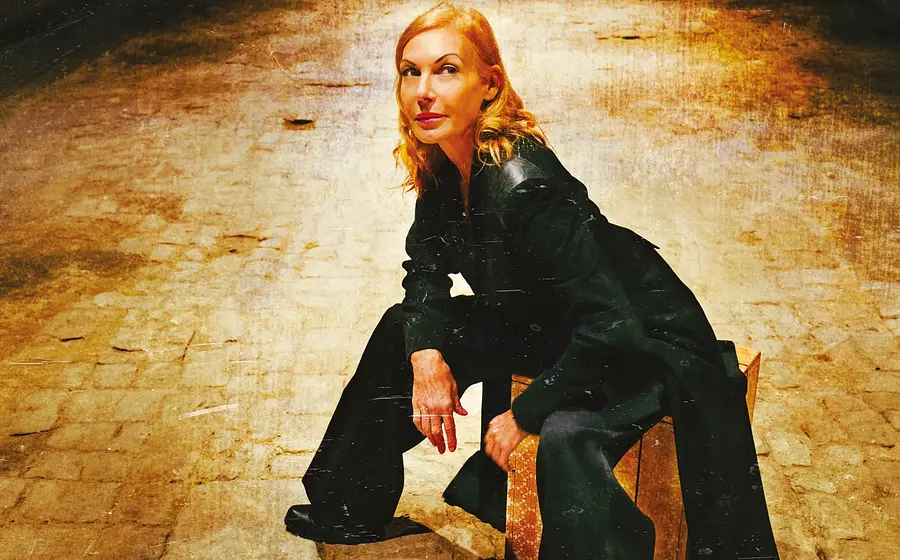 Ute Lemper lächelnd nach oben blickend, in dunkler Kleidung, sitzt auf einem Holzhocker auf gepflastertem Boden in warm beleuchtetem Hintergrund in Gold- und Brauntönen