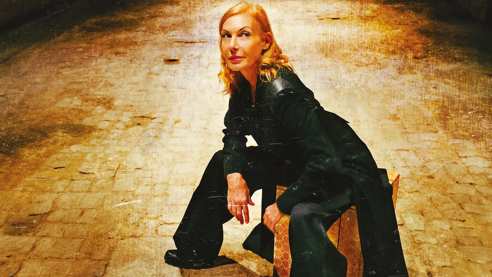 Ute Lemper lächelnd nach oben blickend, in dunkler Kleidung, sitzt auf einem Holzhocker auf gepflastertem Boden in warm beleuchtetem Hintergrund in Gold- und Brauntönen