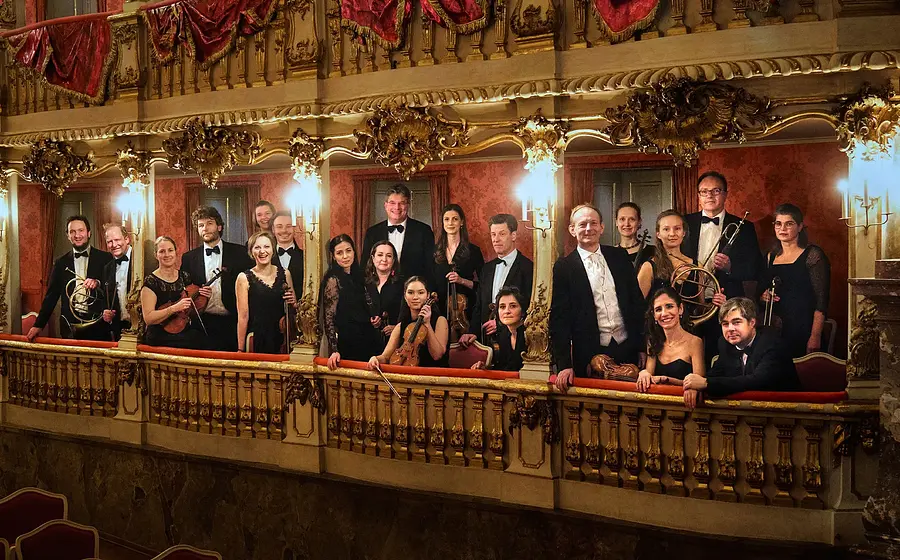 Gruppenfoto eines Orchesters auf einem reich verzierten Theaterbalkon mit goldenen Ornamenten und roten Vorhängen mehrere Personen in schwarzer Konzertkleidung halten Instrumente in einem historischen Saal