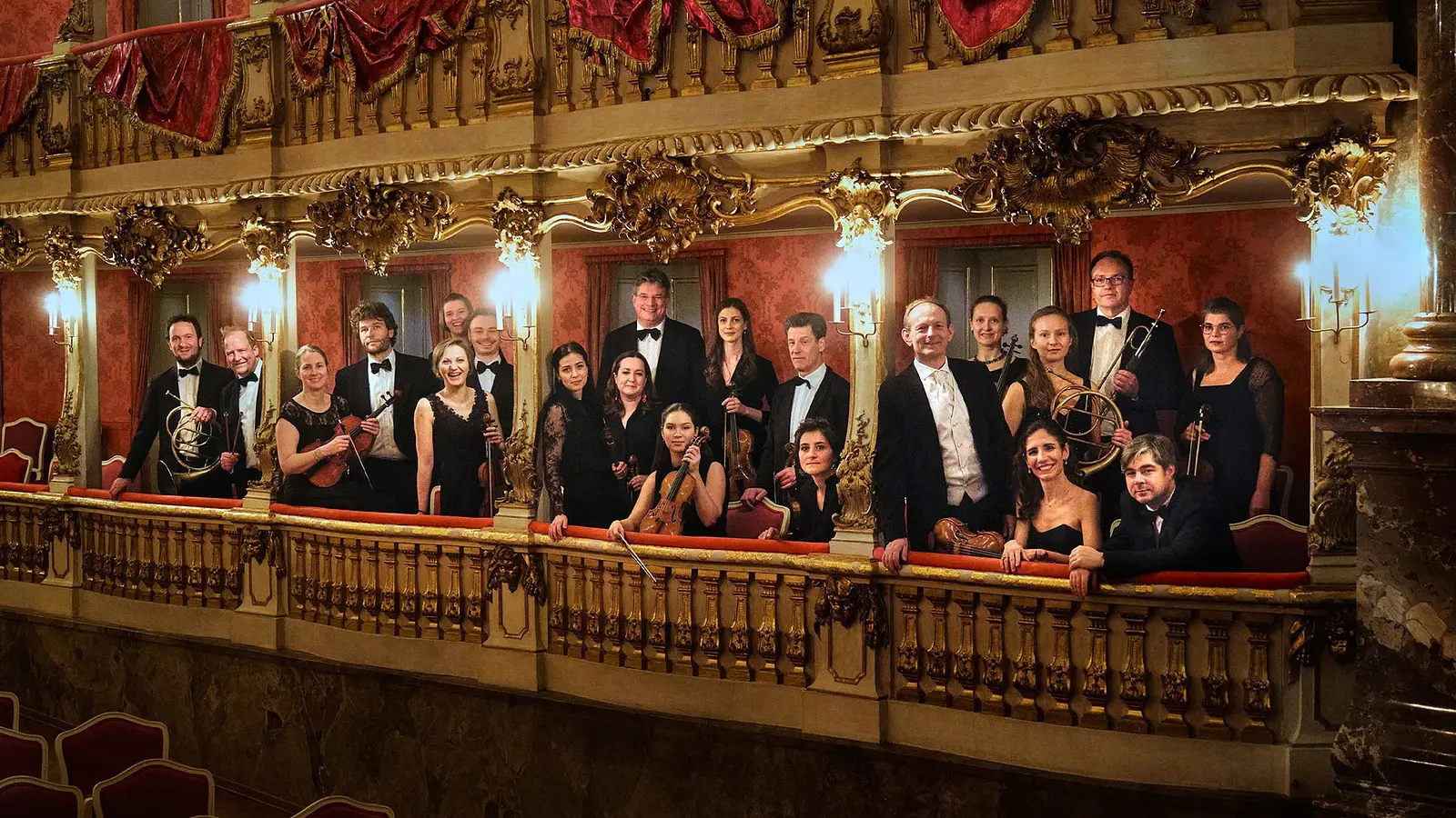 Gruppenfoto eines Orchesters auf einem reich verzierten Theaterbalkon mit goldenen Ornamenten und roten Vorhängen mehrere Personen in schwarzer Konzertkleidung halten Instrumente in einem historischen Saal