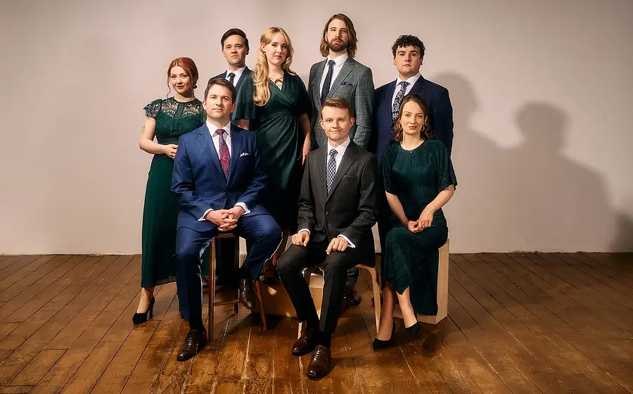 VOCES8 mit acht Sänger:innen in eleganter Kleidung, teilweise sitzend und stehend angeordnet auf einer Holzfläche vor hellem Hintergrund in einer Studioaufnahme.