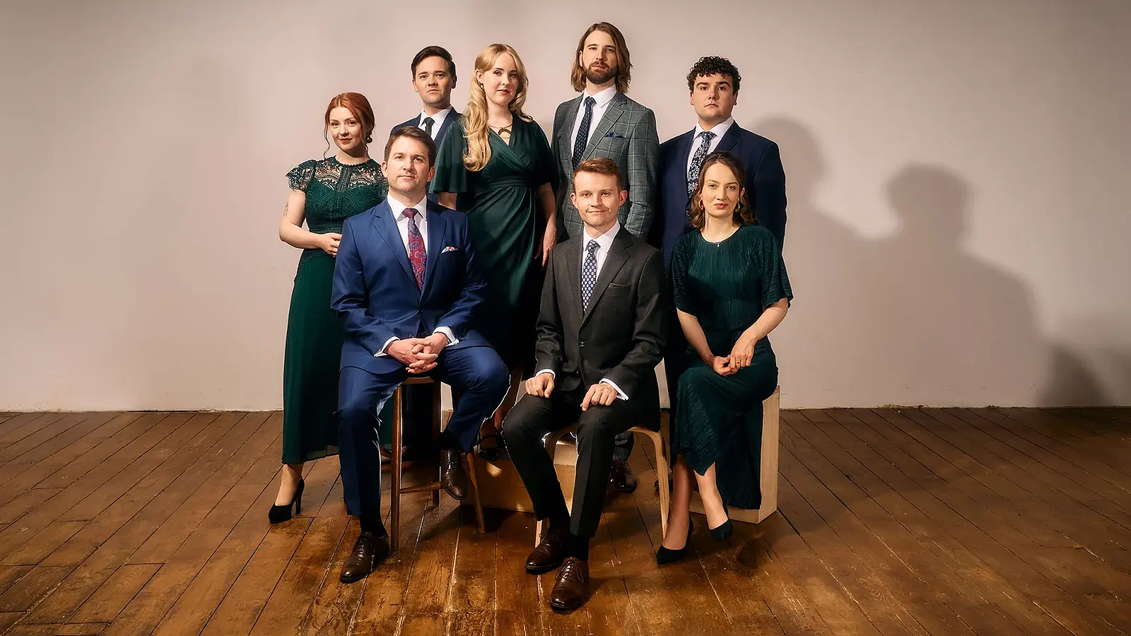 VOCES8 mit acht Sänger:innen in eleganter Kleidung, teilweise sitzend und stehend angeordnet auf einer Holzfläche vor hellem Hintergrund in einer Studioaufnahme.