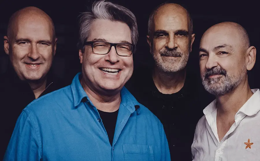Bandfoto von Colorbox mit vier Bandmitgliedern vor dunklem Hintergrund, eine Person im blauen Hemd im Vordergrund, die weiteren in dunkler und weißer Kleidung dahinter angeordnet.