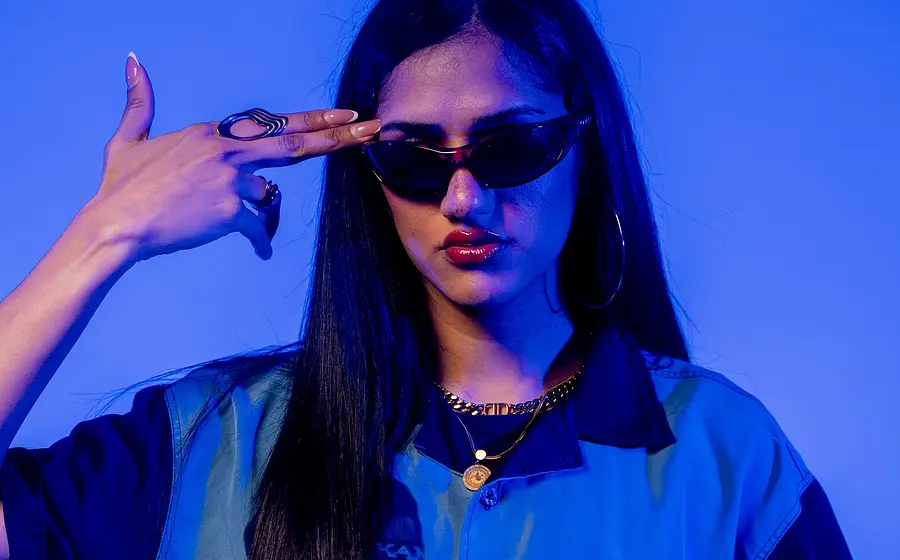 Porträtfoto von Wa22ermann mit langen dunklen Haaren, schmaler Sonnenbrille und blauem Hemd vor blau violett beleuchtetem Hintergrund, die Hand in Form einer Pistole an die Schläfe haltend.
