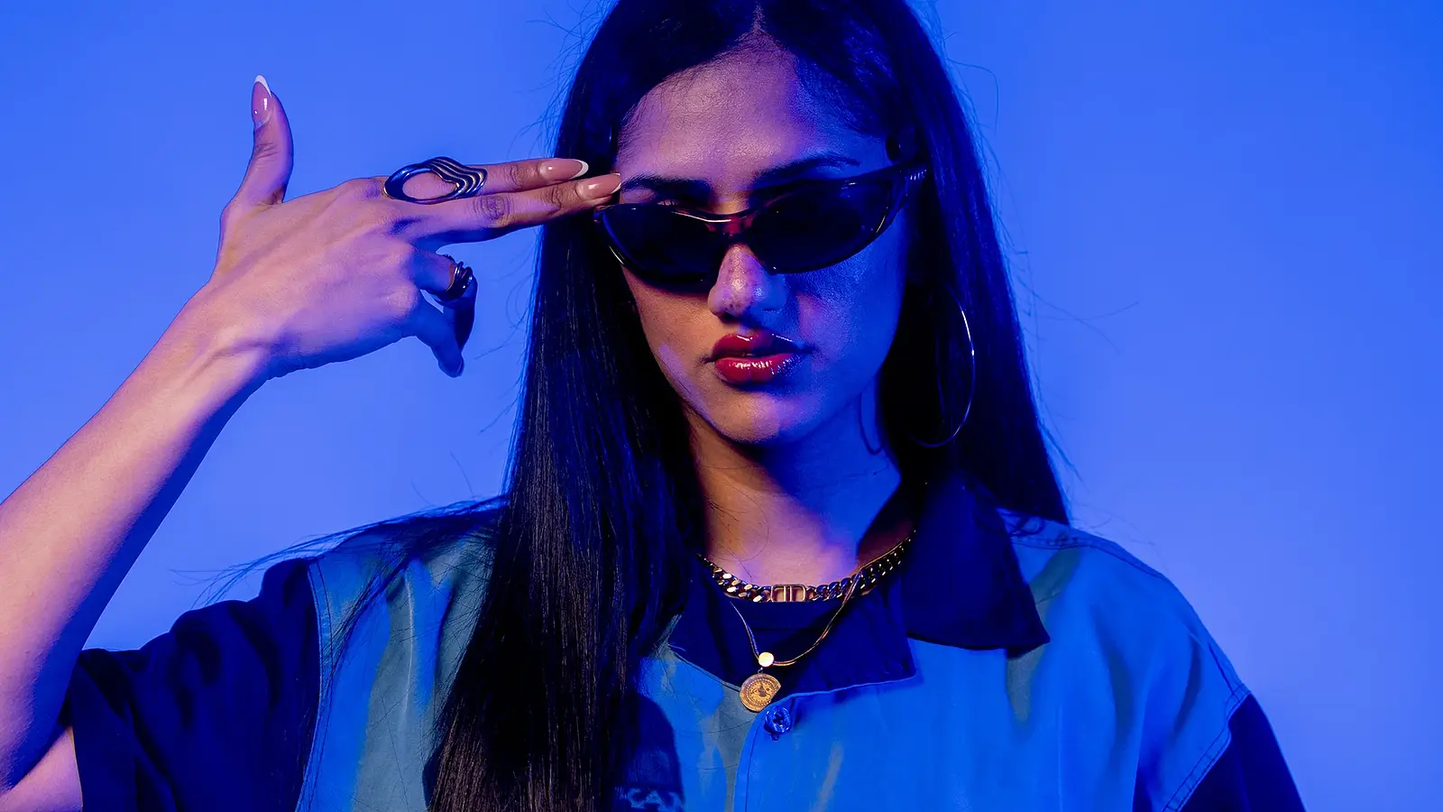 Porträtfoto von Wa22ermann mit langen dunklen Haaren, schmaler Sonnenbrille und blauem Hemd vor blau violett beleuchtetem Hintergrund, die Hand in Form einer Pistole an die Schläfe haltend.