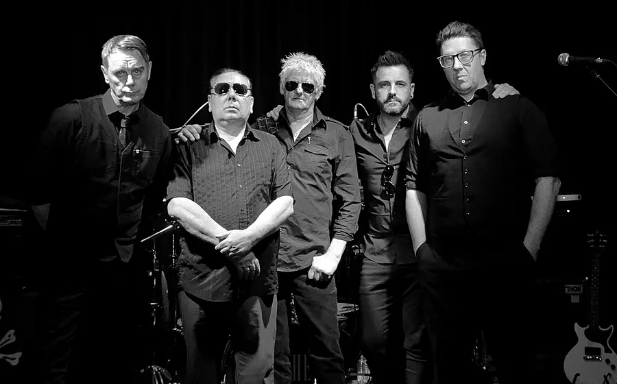 Bandfoto von The Godfathers mit fünf Bandmitgliedern in dunkler Kleidung auf einer Bühne vor schwarzem Hintergrund, zwei Personen mit Sonnenbrillen im Zentrum, umgeben von Mikrofonständern, Schlagzeug und E-Gitarre in Schwarzweißaufnahme.