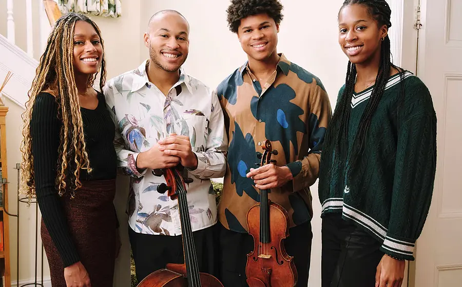 Die vier Mitglieder der Kanneh Masons lachend in bunter Kleidung, zwei Personen mit Instrumenten, vor hellem Hintergrund