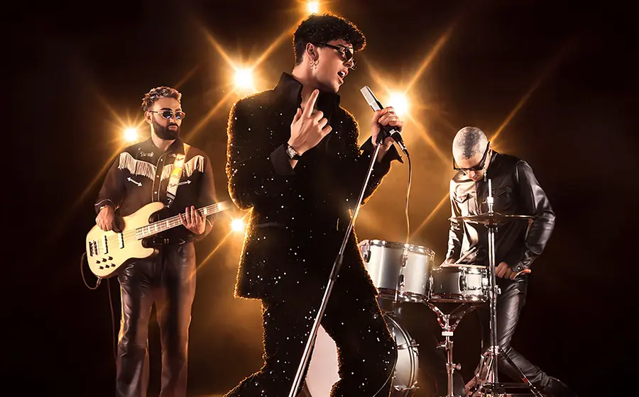 Bandfoto von The Kolors mit drei Musikern vor dunklem Hintergrund und warmen Scheinwerfern, eine Person singt am Mikrofon in glitzerndem schwarzem Outfit, begleitet von E-Bass und Schlagzeug