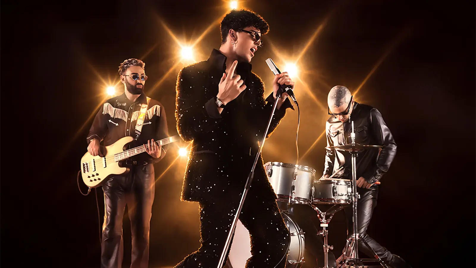 Bandfoto von The Kolors mit drei Musikern vor dunklem Hintergrund und warmen Scheinwerfern, eine Person singt am Mikrofon in glitzerndem schwarzem Outfit, begleitet von E-Bass und Schlagzeug