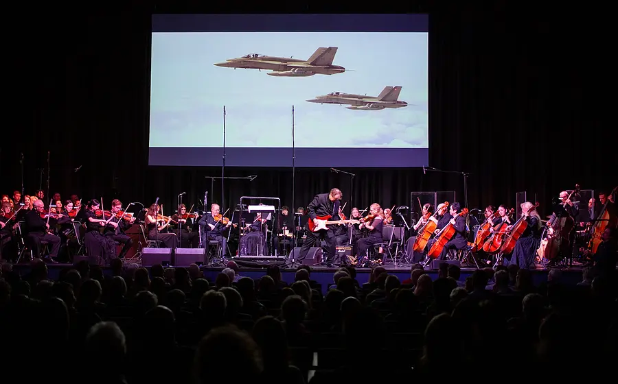 Bühnenfoto eines Orchesters mit Streichern und Gitarrist auf einer Konzertbühne vor Publikum, darüber eine große Leinwand mit der Projektion zweier Kampfjets über den Wolken zur Veranstaltung The Magic of Hans Zimmer