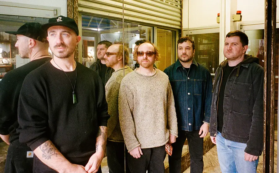 Bandfoto von The Menzingers mit vier Bandmitgliedern in Freizeitkleidung vor einer Schaufensterfront mit Spiegelungen, darunter eine Person mit schwarzer Kappe, eine mit Sonnenbrille und drei weitere in Pullovern und Jacken