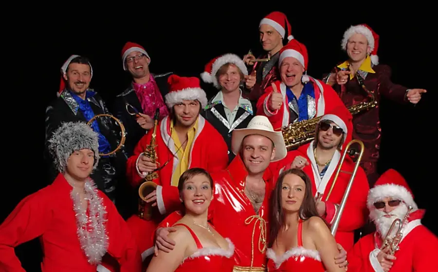 Foto von The Presleys Family in roten Weihnachtskostümen und Weihnachtsmützen vor rot schwarzem Strahlenhintergrund. Die große Musikgruppe posiert mit Saxofon, Posaune, Trompete, Tamburin und weiteren Instrumenten für ein festliches Weihnachtsmotiv.
