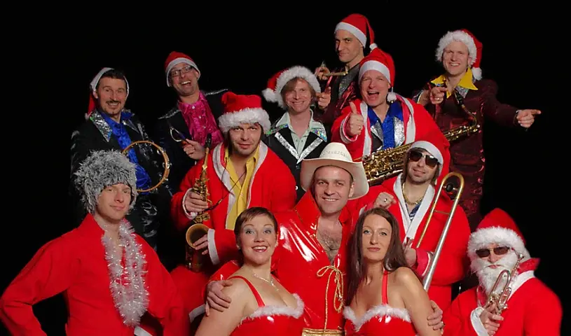 Foto von The Presleys Family in roten Weihnachtskostümen und Weihnachtsmützen vor rot schwarzem Strahlenhintergrund. Die große Musikgruppe posiert mit Saxofon, Posaune, Trompete, Tamburin und weiteren Instrumenten für ein festliches Weihnachtsmotiv.