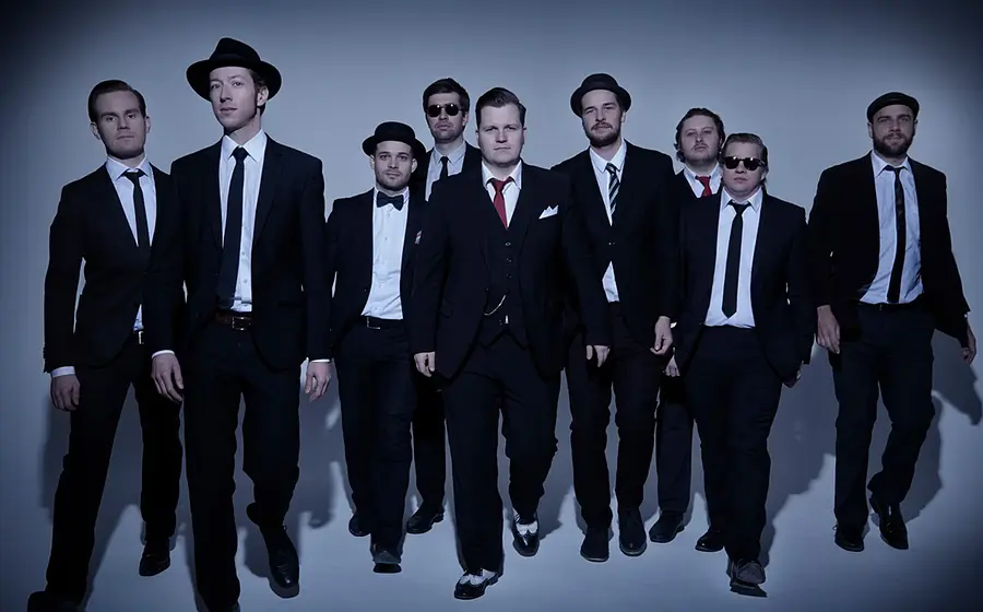 Bandfoto von The Sweet Simones mit neun Personen in dunklen Anzügen und Krawatten, die vor hellem Studiohintergrund auf die Kamera zugehen, einige mit Hut oder Sonnenbrille, eine Person mittig mit roter Krawatte.