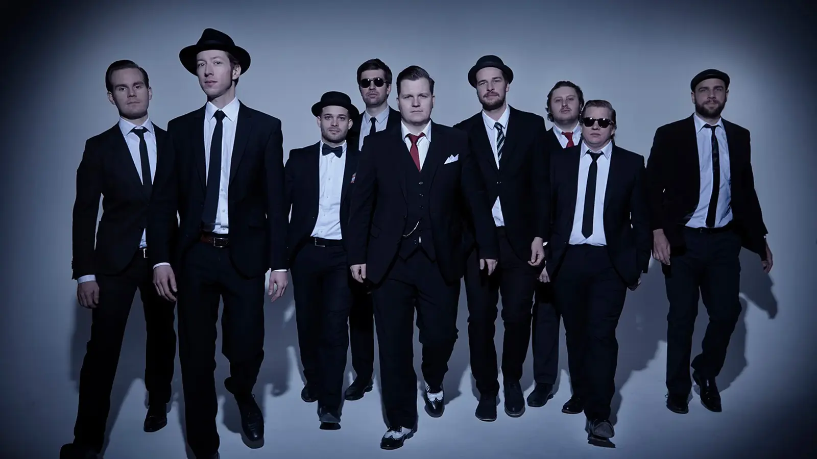 Bandfoto von The Sweet Simones mit neun Personen in dunklen Anzügen und Krawatten, die vor hellem Studiohintergrund auf die Kamera zugehen, einige mit Hut oder Sonnenbrille, eine Person mittig mit roter Krawatte.