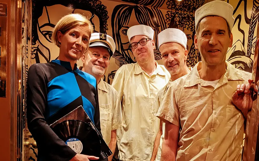 Fünf Mitglieder der Band The Ukelites stehen in einem Aufzug. Eine Person trägt ein blau-schwarzes Outfit und hält eine Schallplatte, vier Personen tragen hellbeige Kleidung mit weißen Mützen. Der Aufzug ist in warmen Braun- und Goldtönen gestaltet und mit großflächigen schwarzen Illustrationen an den Wänden dekoriert.