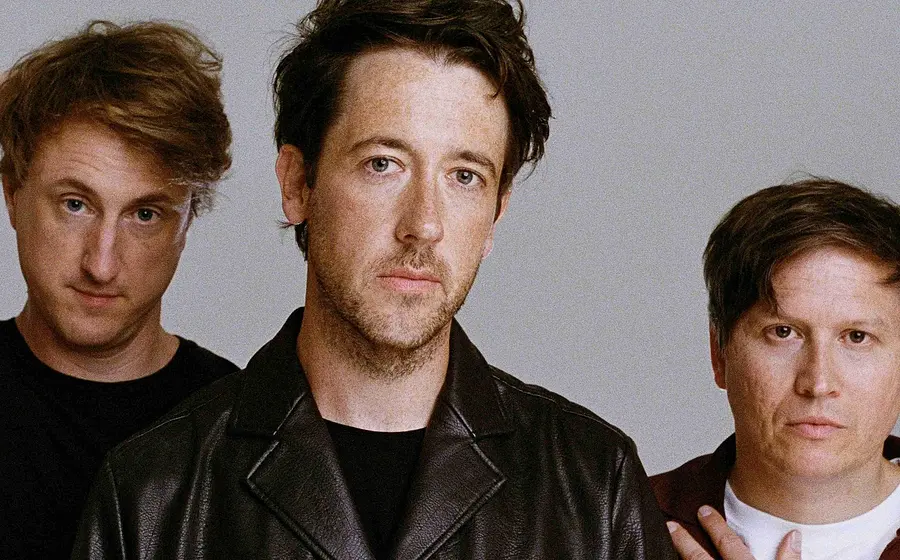Bandfoto von The Wombats mit drei Bandmitgliedern vor hellem Hintergrund, die mittlere Person in schwarzer Lederjacke, flankiert von zwei Personen in dunkler Freizeitkleidung, eine mit aufgelegter Hand am Kopf und eine mit der Hand auf der Schulter.
