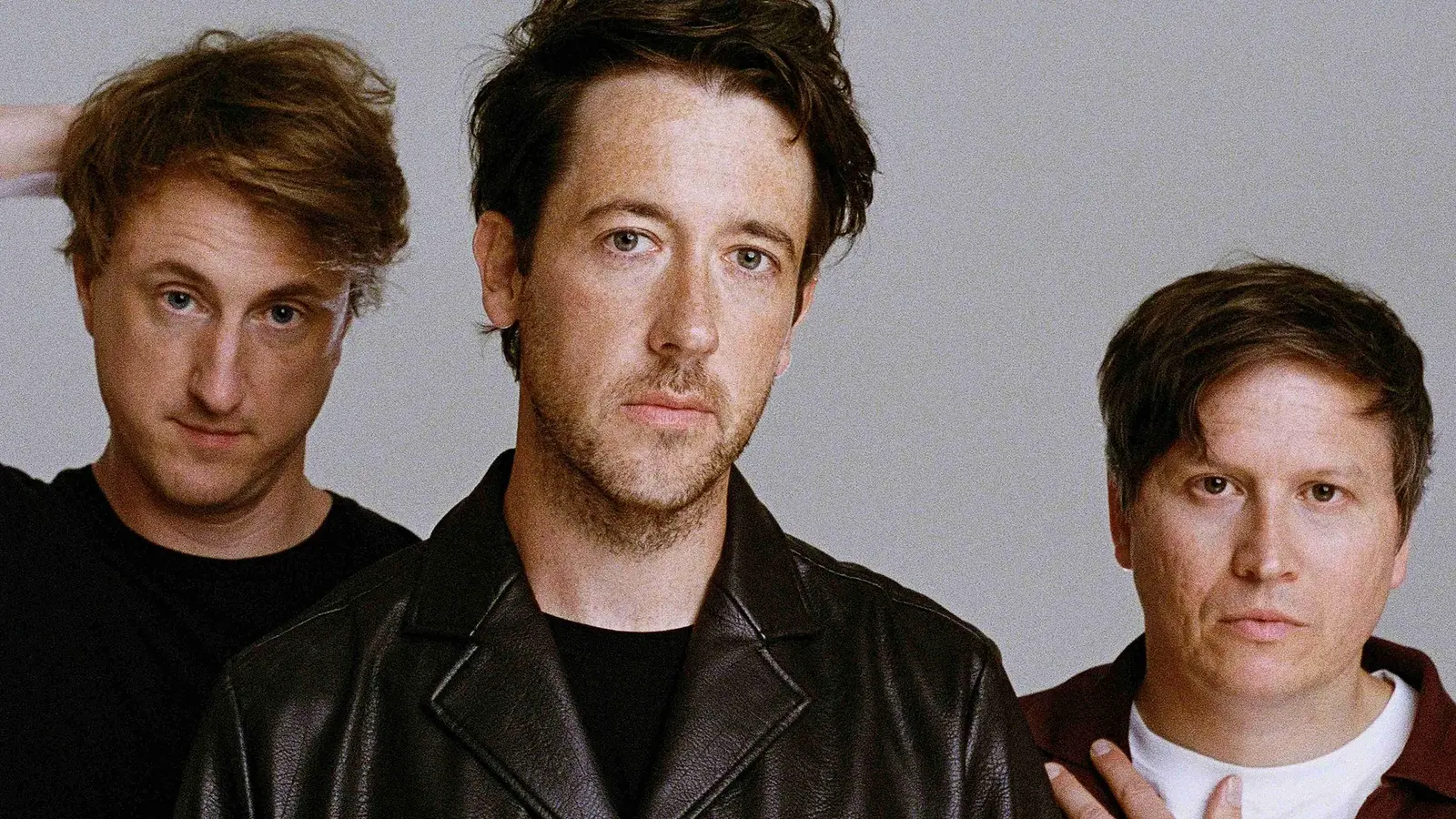 Bandfoto von The Wombats mit drei Bandmitgliedern vor hellem Hintergrund, die mittlere Person in schwarzer Lederjacke, flankiert von zwei Personen in dunkler Freizeitkleidung, eine mit aufgelegter Hand am Kopf und eine mit der Hand auf der Schulter.