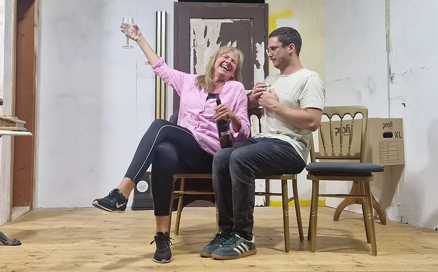 Theaterszene mit zwei sitzenden Personen auf einer Bühne vor einer Wohnungs-Kulisse.