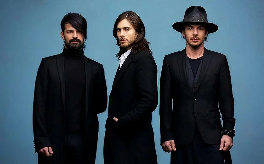 Drei Mitglieder der Band **Thirty Seconds To Mars** stehen nebeneinander vor einem blauen Hintergrund, gekleidet in schwarze, elegante Outfits; einer trägt einen Hut, die anderen haben längeres Haar und einen ernsten Blick in die Kamera.