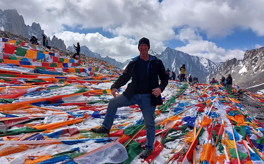 Thomas Bauer steht zwischen bunten Gebetsfahnenauf der Passhoehe Doelma La auf 5600m. Im Hintergrund sind weitere Besucherinnen und Besucher
