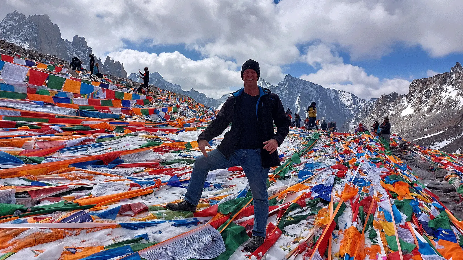 Thomas Bauer steht zwischen bunten Gebetsfahnenauf der Passhoehe Doelma La auf 5600m. Im Hintergrund sind weitere Besucherinnen und Besucher