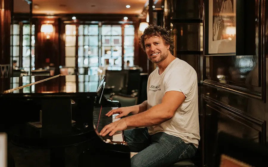 Thomas Bergersen sitzt in weißem Shirt und Jeans an einem Piano und blickt lächelnd in die Kamera, im Hintergrund ein stimmungsvoll beleuchteter Raum mit dunkler Holzausstattung