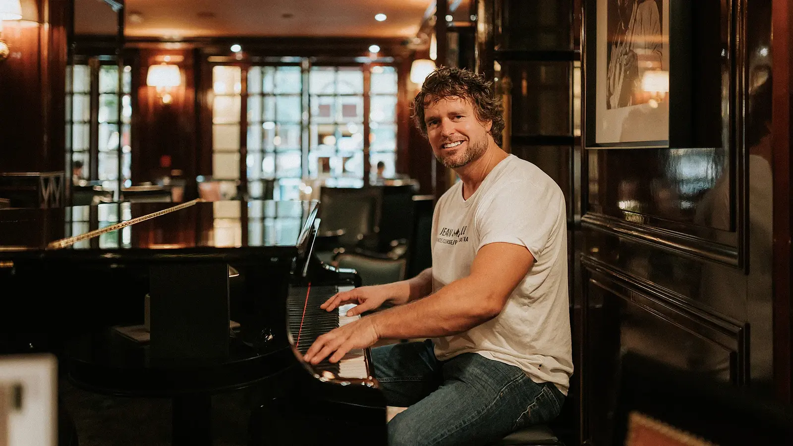 Thomas Bergersen sitzt in weißem Shirt und Jeans an einem Piano und blickt lächelnd in die Kamera, im Hintergrund ein stimmungsvoll beleuchteter Raum mit dunkler Holzausstattung