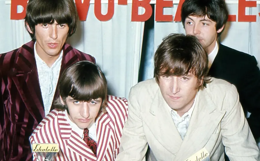 Historisches Pressefoto der Beatles in auffälligen Anzügen vor dem Schriftzug Beatles. Zwei Personen stehen im Hintergrund, während zwei weitere Personen im Vordergrund nach vorne gebeugt in die Kamera blicken.