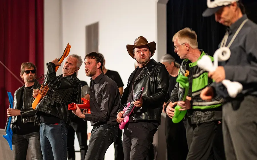 Mehrere Personen stehen auf einer Bühne und spielen mit aufblasbaren Spielzeuginstrumenten. Einige tragen Kostüme, darunter eine Cowboyhut- und Lederjackenkombination