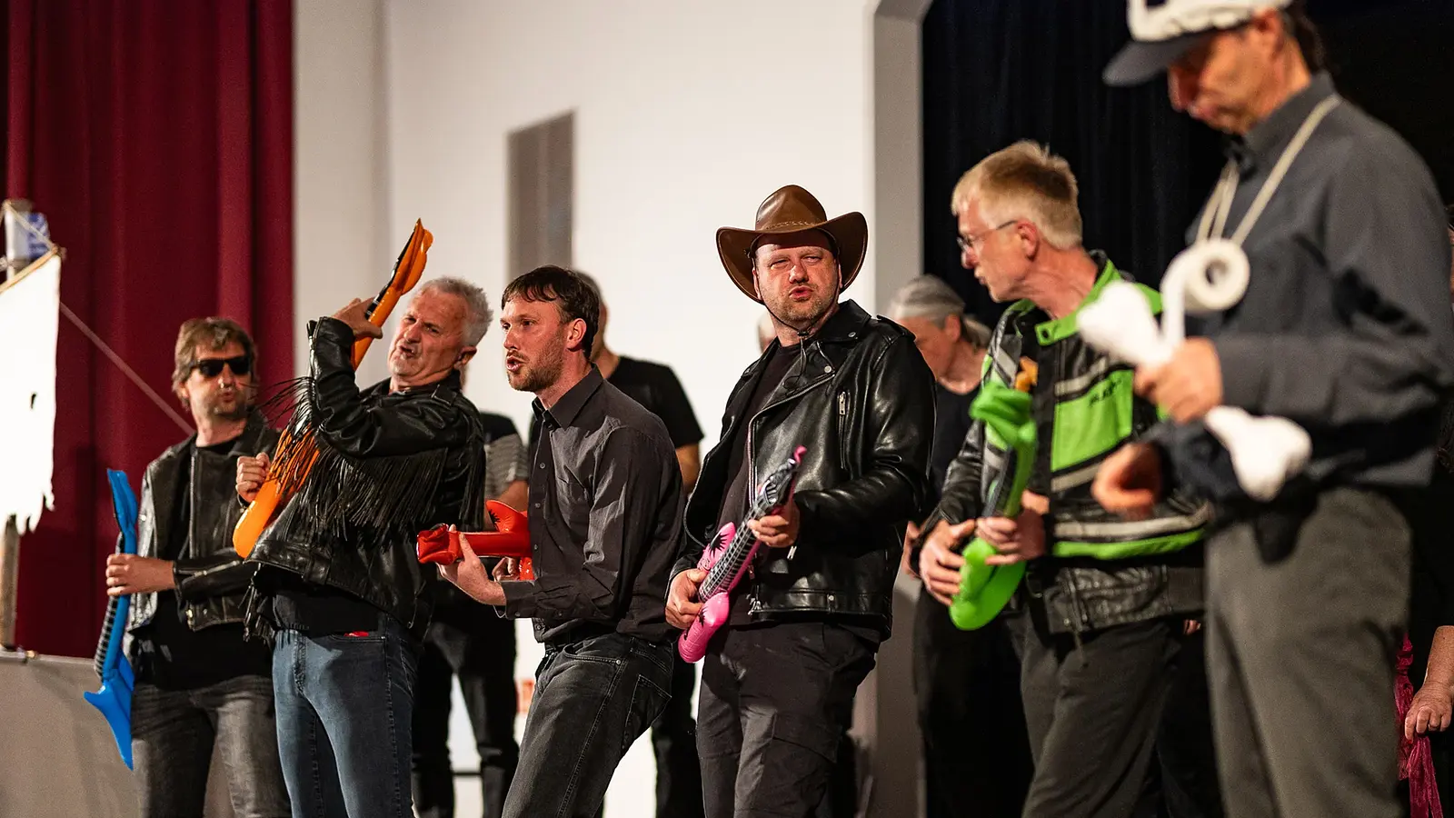 Mehrere Personen stehen auf einer Bühne und spielen mit aufblasbaren Spielzeuginstrumenten. Einige tragen Kostüme, darunter eine Cowboyhut- und Lederjackenkombination