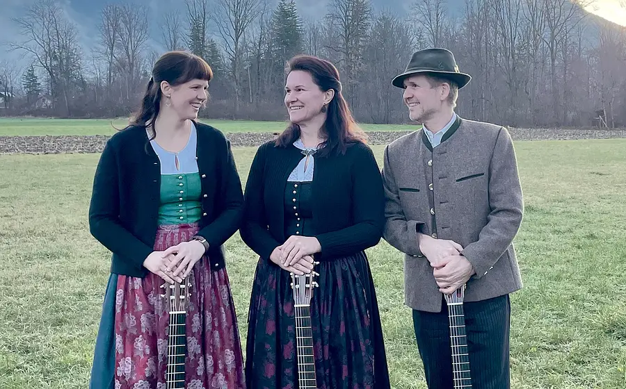 Drei Personen in Tracht mit je einer akustischen Gitarre auf einer Wiese. Zwei Frauen in Dirndln in Grün-, Rosa- und Schwarztönen, ein Mann mit graubrauner Trachtenjacke und Hut. Hintergrund mit Wiese, kahlen Bäumen und Bergen