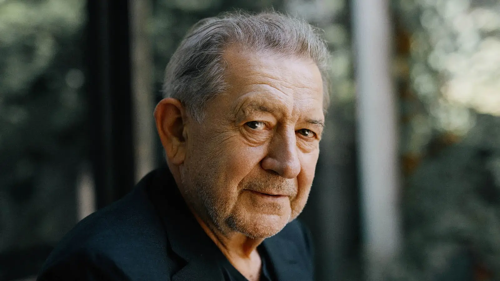 Portrait von André Jung in einem schwarzen Sakko seitlich in die Kamera blickend, der Hintergrund verschwommen