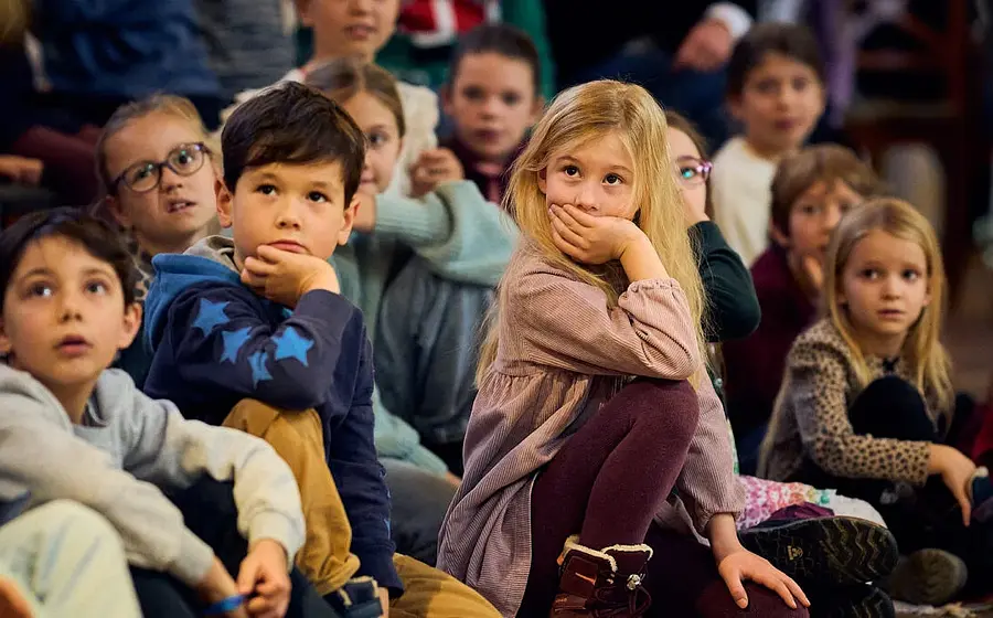 Foto mehrerer Kinder, die auf dem Boden sitzen und einer Aufführung folgen, im Vordergrund ein Kind im rosafarbenen Kleid sowie ein Kind im blauen Pullover mit Sternenmuster, umgeben von weiteren Kindern in einem Veranstaltungsraum.