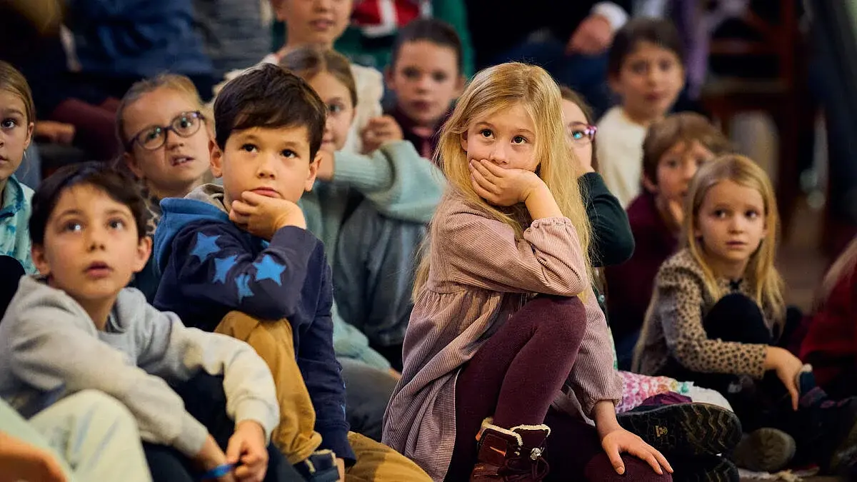 Foto mehrerer Kinder, die auf dem Boden sitzen und einer Aufführung folgen, im Vordergrund ein Kind im rosafarbenen Kleid sowie ein Kind im blauen Pullover mit Sternenmuster, umgeben von weiteren Kindern in einem Veranstaltungsraum.
