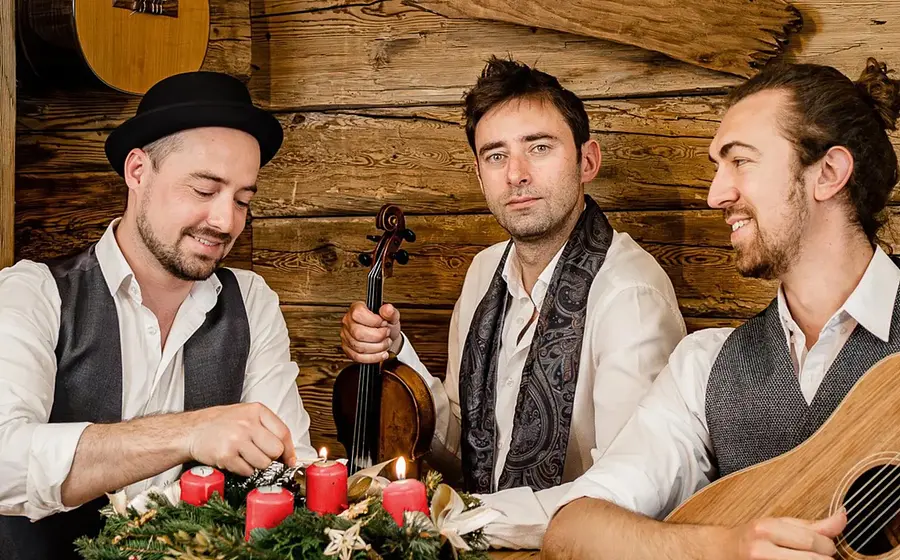 Drei Musiker in weißer und dunkler Kleidung sitzen und stehen in einer Holzhütte; einer zündet Kerzen an einem Adventskranz, während die anderen mit Gitarre und Violine posieren.