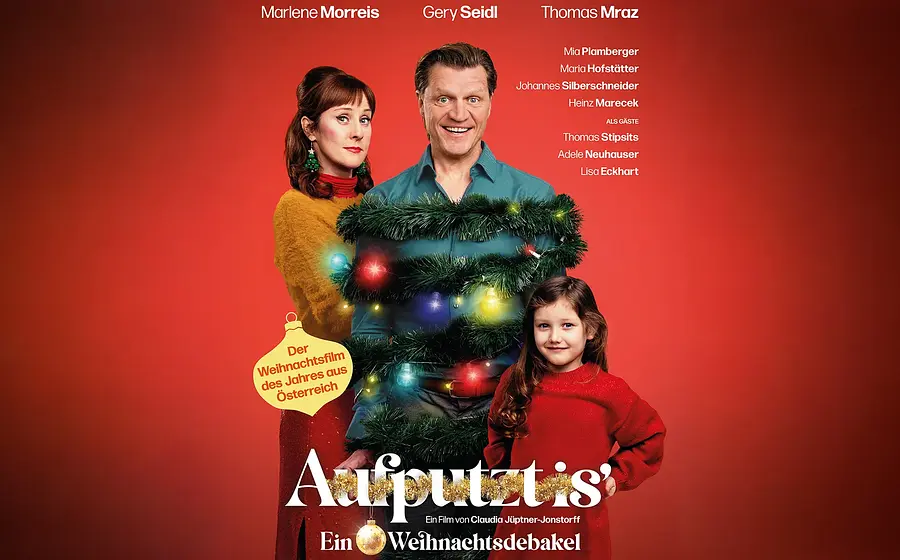 Plakat Aufputzt mit drei Personen vor rotem Hintergrund, eine Figur mit Weihnachtsgirlande und bunten Lichtern geschmückt in der Mitte, daneben zwei weitere Personen in farbiger Kleidung, darunter großer Titel Aufputzt und Zusatz Ein Weihnachtsdebakel