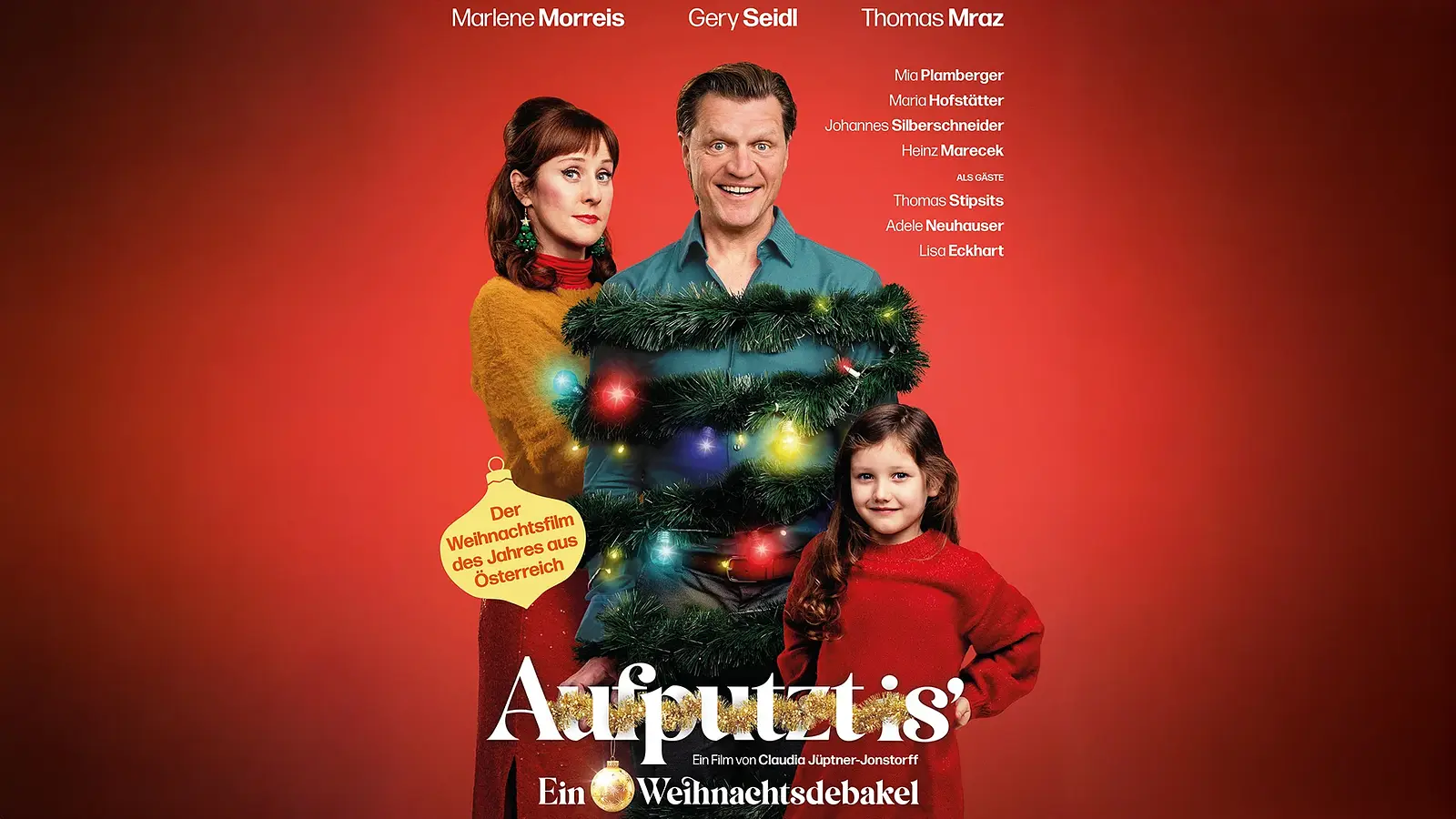 Plakat Aufputzt mit drei Personen vor rotem Hintergrund, eine Figur mit Weihnachtsgirlande und bunten Lichtern geschmückt in der Mitte, daneben zwei weitere Personen in farbiger Kleidung, darunter großer Titel Aufputzt und Zusatz Ein Weihnachtsdebakel