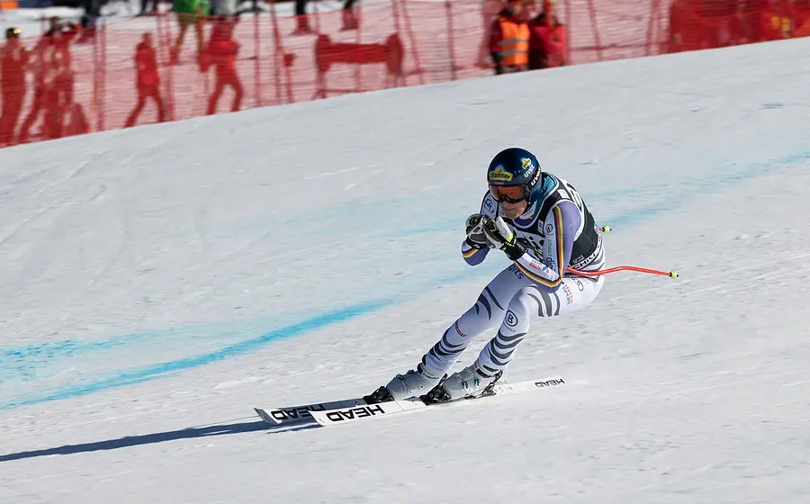 Foto eines Skirennläufers in weiß grauem Rennanzug und blauem Helm bei hoher Geschwindigkeit auf einer präparierten Skipiste, in aerodynamischer Abfahrtshaltung vor rotem Sicherheitsnetz und Zuschauerbereich im Hintergrund