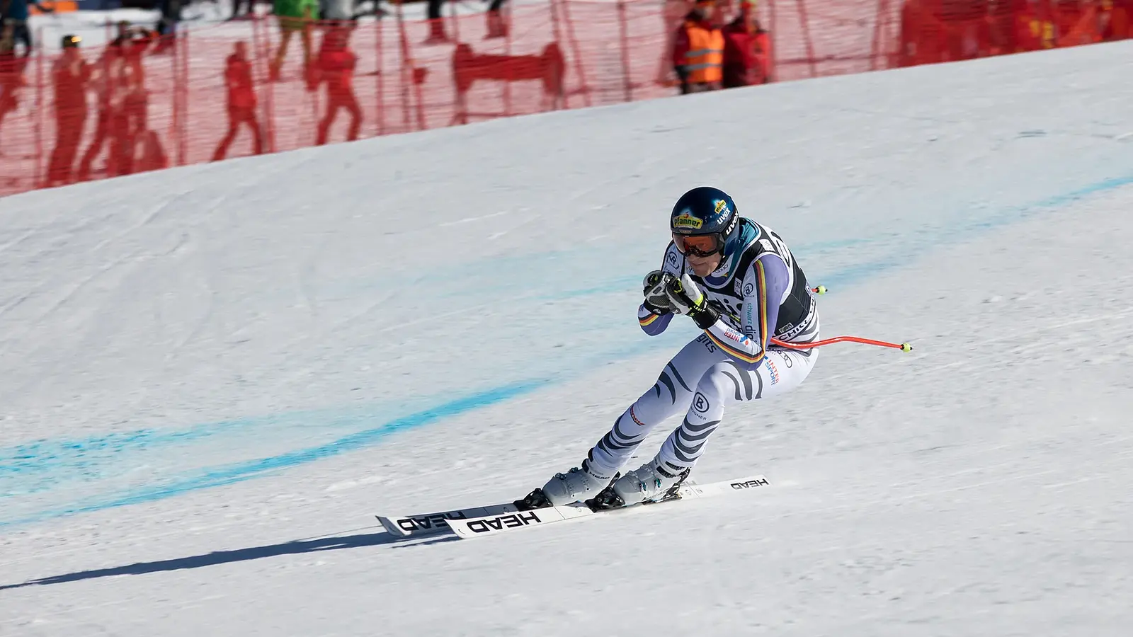 Foto eines Skirennläufers in weiß grauem Rennanzug und blauem Helm bei hoher Geschwindigkeit auf einer präparierten Skipiste, in aerodynamischer Abfahrtshaltung vor rotem Sicherheitsnetz und Zuschauerbereich im Hintergrund
