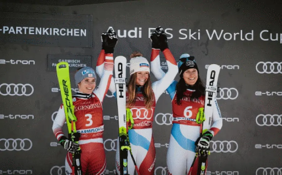 Foto der Siegerinnen eines Ski-Weltcuprennens bei der Siegerehrung in Garmisch-Partenkirchen, drei Athletinnen in Rennanzügen halten ihre Skier und stehen vor einer Sponsorenwand mit Audi-Logos, die Person in der Mitte hebt die Arme nach oben