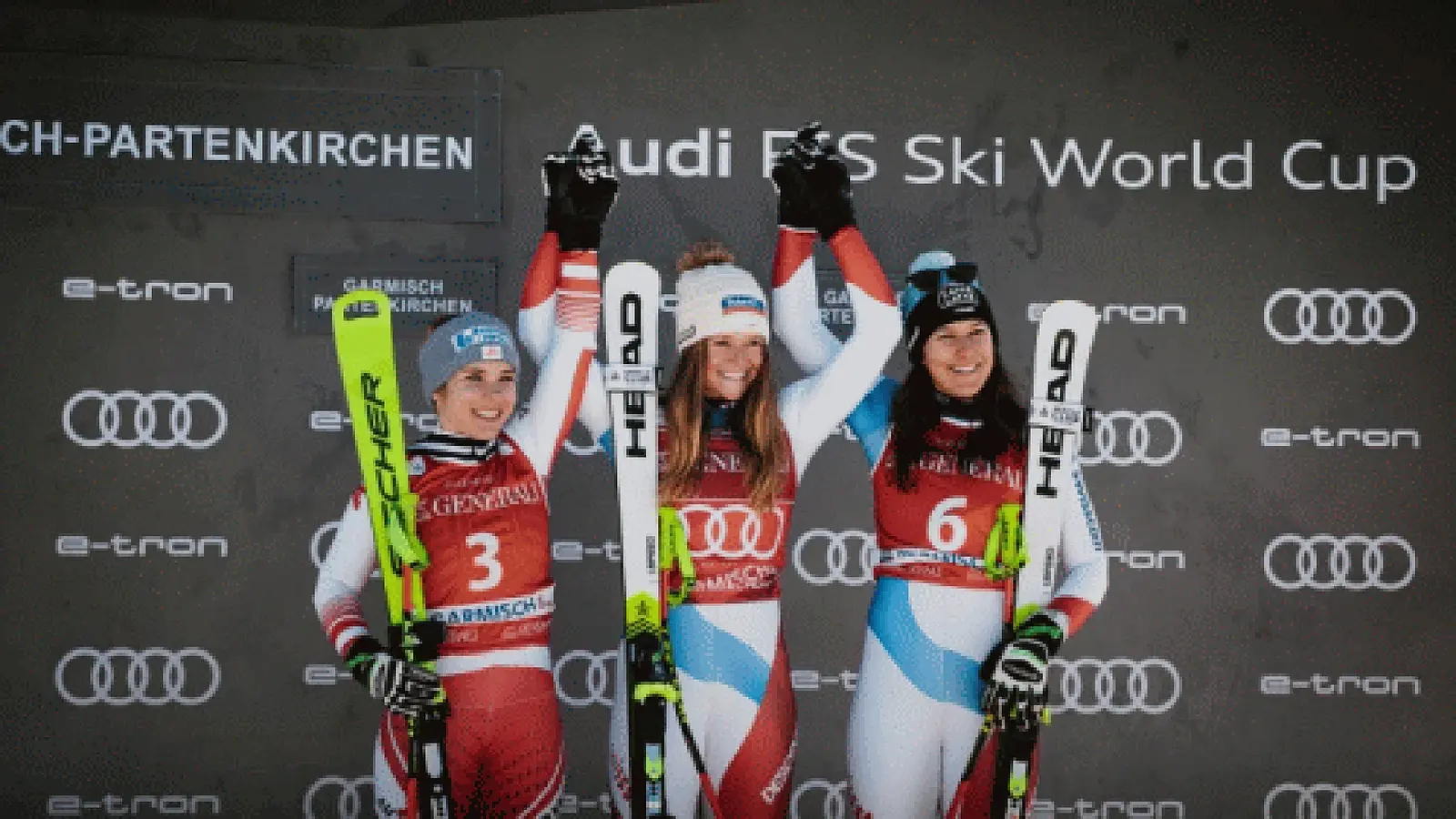 Foto der Siegerinnen eines Ski-Weltcuprennens bei der Siegerehrung in Garmisch-Partenkirchen, drei Athletinnen in Rennanzügen halten ihre Skier und stehen vor einer Sponsorenwand mit Audi-Logos, die Person in der Mitte hebt die Arme nach oben