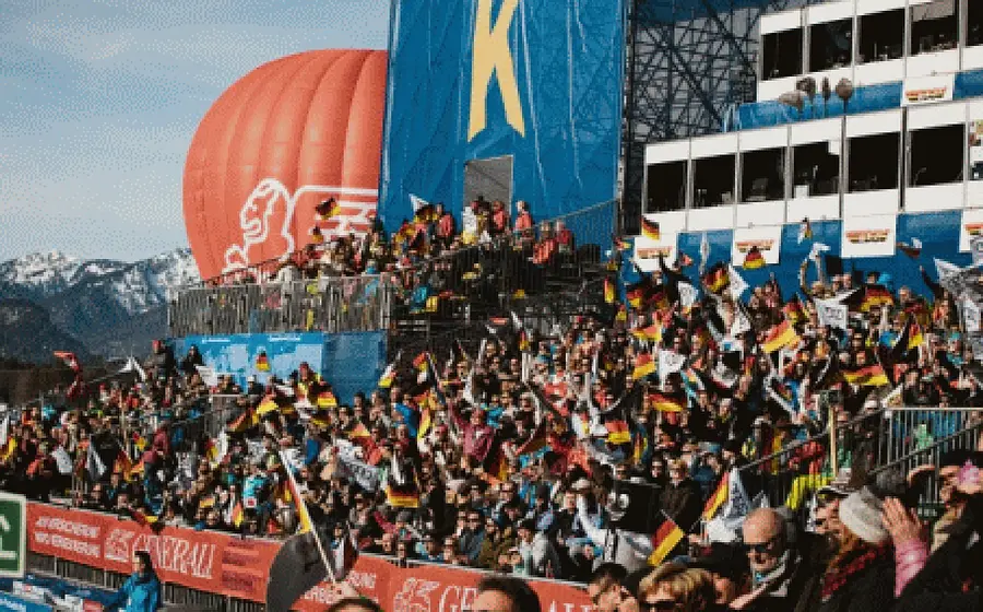 Foto vom Ski-Weltcup in Garmisch-Partenkirchen mit voll besetzten Zuschauertribünen, zahlreichen Deutschlandfahnen im Publikum, großem Kandahar-Ballon und verschneiter Bergkulisse im Hintergrund