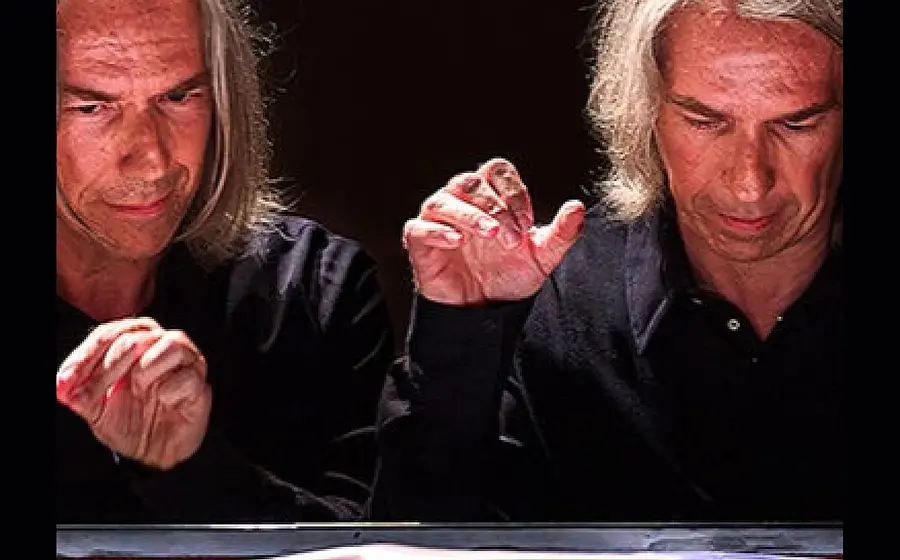 Porträtfoto eines Pianisten mit schulterlangem grauem Haar am geöffneten Konzertflügel, dessen Gesicht und Hände in einer Doppelbelichtung zweimal dargestellt sind, vor dunklem Hintergrund und beleuchteter Klaviatur.