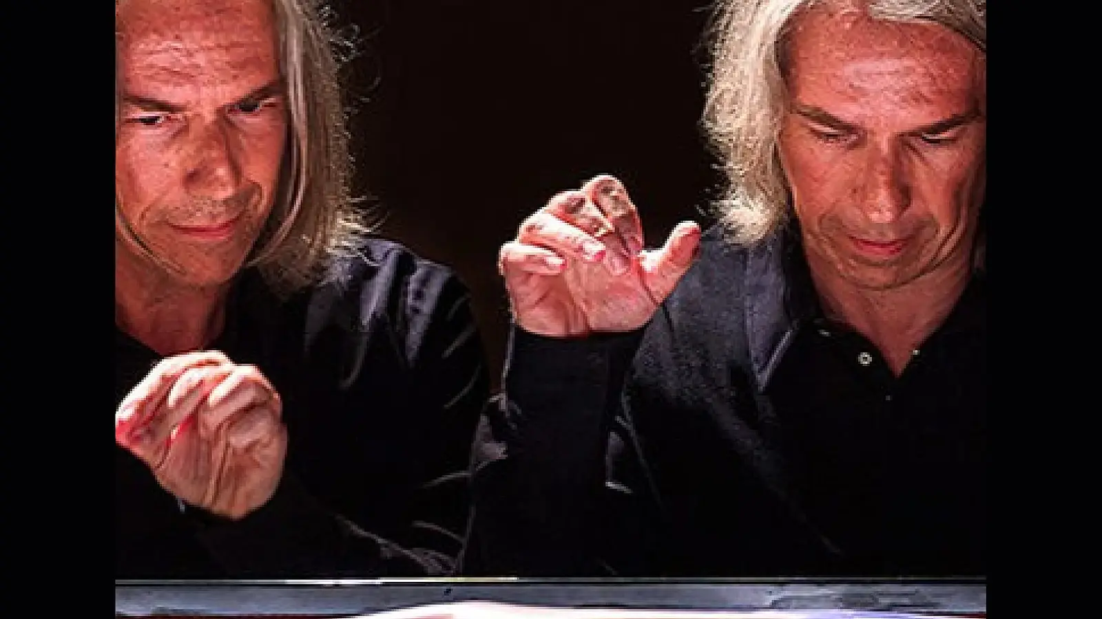 Porträtfoto eines Pianisten mit schulterlangem grauem Haar am geöffneten Konzertflügel, dessen Gesicht und Hände in einer Doppelbelichtung zweimal dargestellt sind, vor dunklem Hintergrund und beleuchteter Klaviatur.
