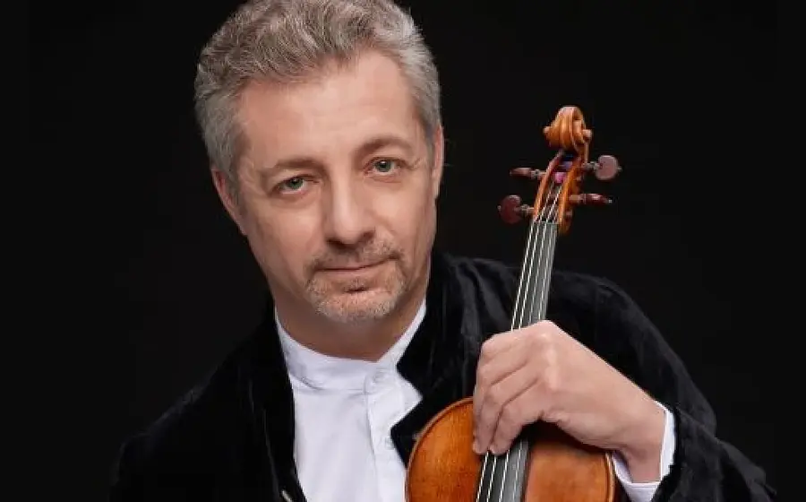 Porträtfoto eines Violinisten in schwarzem Konzertsakko und weißem Hemd vor schwarzem Hintergrund, der eine Violine in der Hand hält und direkt zur Kamera blickt.