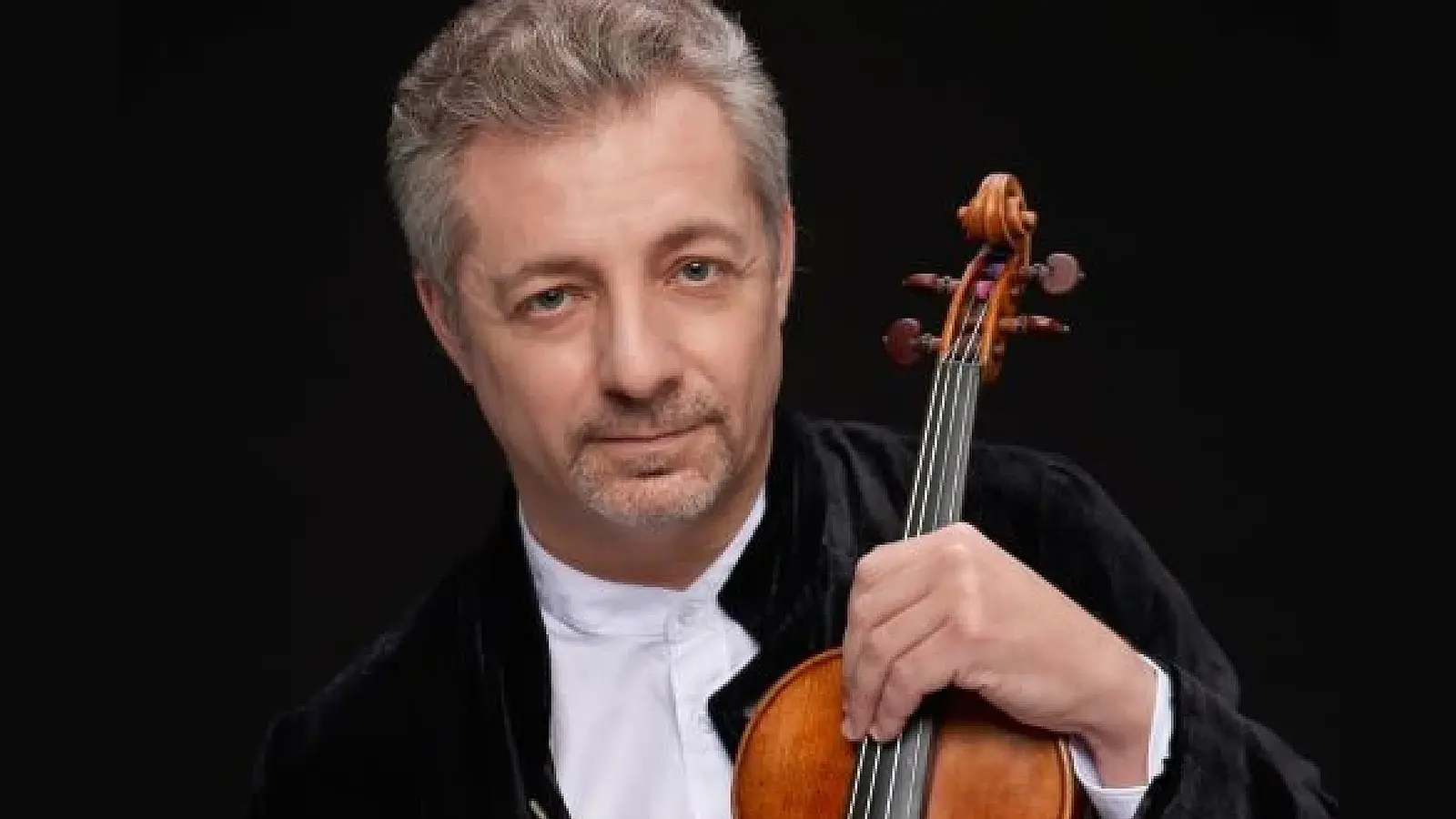 Porträtfoto eines Violinisten in schwarzem Konzertsakko und weißem Hemd vor schwarzem Hintergrund, der eine Violine in der Hand hält und direkt zur Kamera blickt.